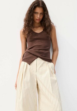 Frau mit langen lockigen Haaren, die ein ärmelloses braunes Oberteil und lockere cremefarbene Hosen mit dünnen vertikalen Streifen trägt, vor einem weißen Hintergrund stehend.
