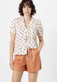Hvit kortermet skjorte med blomstermønster i rødt og grønt, med knytelukking i livet. Kombinert med oransje shorts.