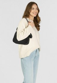 Borsa a tracolla nera in pelle con forma curva, superficie texturizzata e dettaglio della zip. Il modello indossa un maglione crema e jeans azzurri chiari.