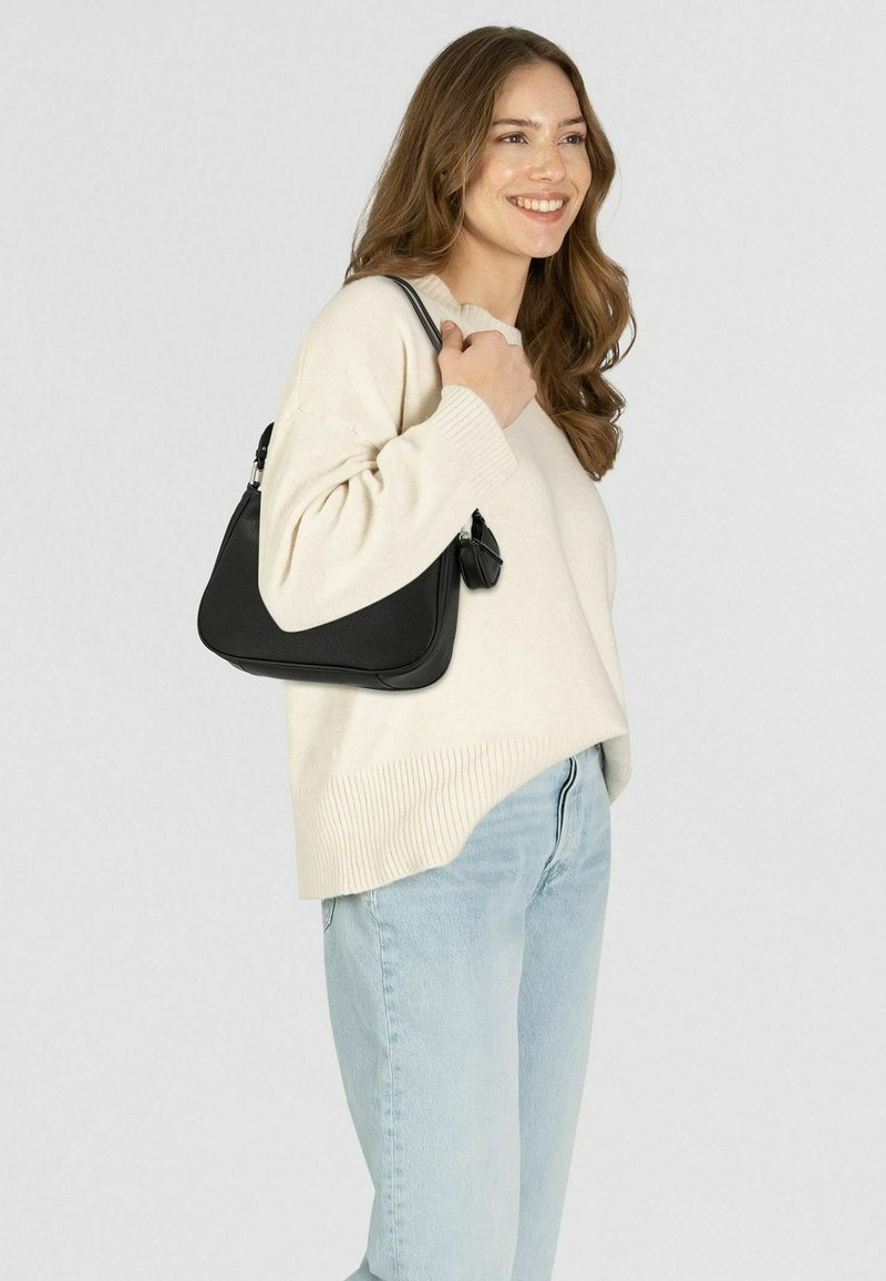 Borsa a tracolla nera in pelle con forma curva, superficie texturizzata e dettaglio della zip. Il modello indossa un maglione crema e jeans azzurri chiari.
