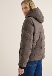 Veste matelassée marron avec capuche, présentant un design matelassé, un tissu texturé et un patch logo sur la manche gauche. Assortie à un jean bleu.
