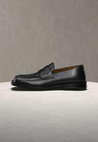 Μαύρο δερμάτινο loafer penny με χαμηλό τακούνι και λεπτομέρεια ραφής, τοποθετημένο σε φωτεινή επιφάνεια μπροστά από ουδέτερο φόντο.