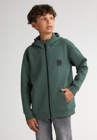 Svetrová mikina s kapucí v tmavě zelené barvě, vyrobená z měkké látky. Obsahuje plný zip, přední kapsy a černou nášivku s logem na hrudi.