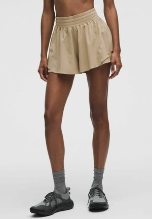 Donna con pantaloncini sportivi beige, calze grigie e scarpe da corsa grigio scuro, in piedi davanti a uno sfondo chiaro e semplice.