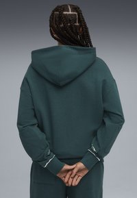Sudadera con capucha en un tono verde azulado oscuro, con un corte relajado, parches acolchados en los codos y costuras decorativas en los puños.