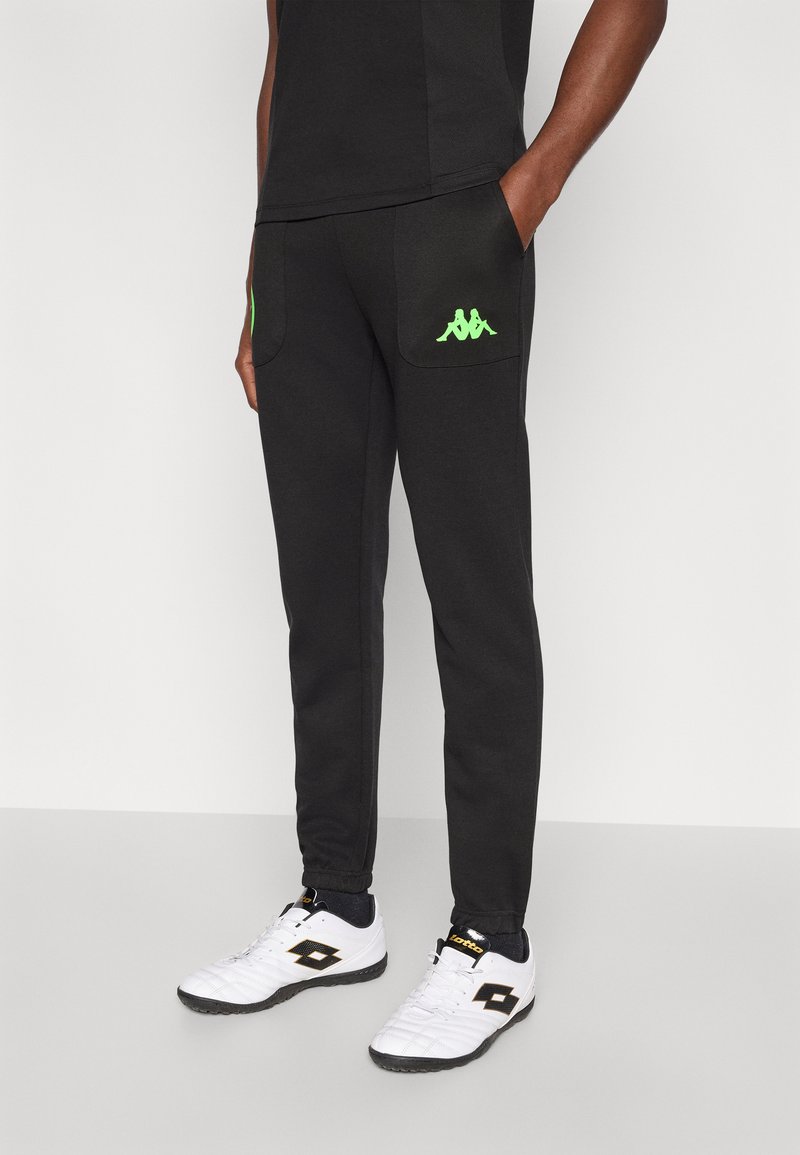 Čierne tepláky s zelenými logami Kappa na vreckách, elastickými lemy a bočnými vreckami, doplnené bielo-čiernymi športovými topánkami.