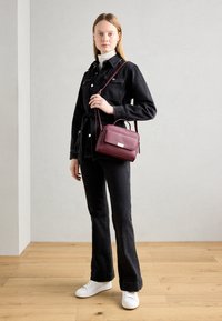 Borsa a tracolla bordeaux con finitura testurizzata, forma rettangolare, hardware dorato. Indossata con un giubbotto di jeans neri e jeans a zampa d'elefante, abbinata a sneaker bianche.