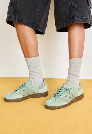 Benendragen met lichtgrijze sokken en mintgroene sneakers met bruine zolen, staand op een okergele vloer, gekleed in zwarte cropped broek.