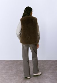 Chaleco de piel sintética marrón sobre un sudadera gris, combinado con pantalones gris holgados y zapatos de color claro. Textura suave sin patrón visible.