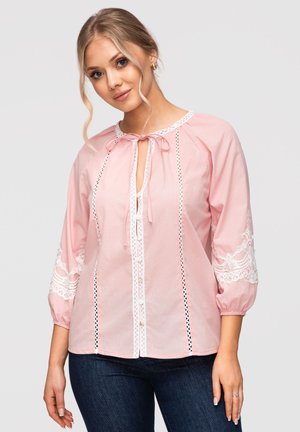 Femme blonde portant un chemisier rose clair avec des détails en dentelle blanche, manches trois-quarts et jean bleu foncé sur un fond uni.