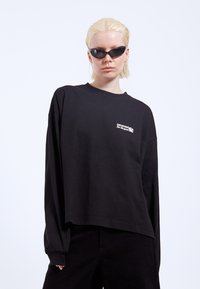 Black long-sleeve T-shirt with a relaxed fit, featuring a small white embroidered logo on the chest and cuffed sleeves.