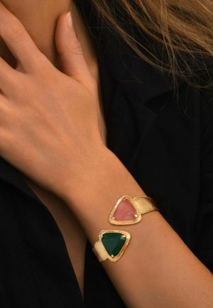 Bracelet manchette en or avec de grandes pierres triangulaires roses et vertes porté au poignet d'une femme contre une manche noire.