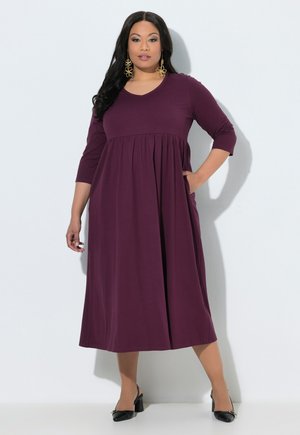Femme aux longs cheveux noirs portant une robe midi violette à manches longues, des talons noirs et de grandes boucles d’oreilles dorées en forme de fleur, debout devant un fond blanc.