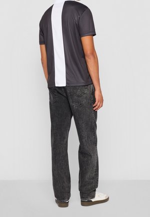 Personne portant un maillot de sport rayé verticalement noir et blanc, un jean noir délavé et des baskets blanches, debout de dos.