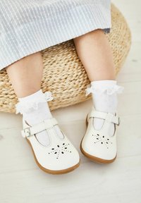 Białe skórzane buty dla małych dzieci z wyciętymi wzorami kwiatowymi, brązowymi gumowymi podeszwami i regulowanymi paskami. W zestawie z białymi skarpetkami z koronkowym wykończeniem.