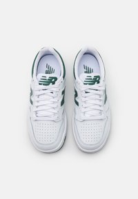 Bílé sportovní boty s zelenými akcenty, s perforovanou špičkou, plochými tkaničkami a logem New Balance na boku a jazyku.