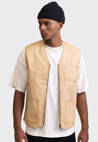 Chaleco de algodón beige con dos bolsillos frontales y cierre de cremallera. Llevado sobre una camiseta blanca, presenta un diseño minimalista y un ajuste casual.