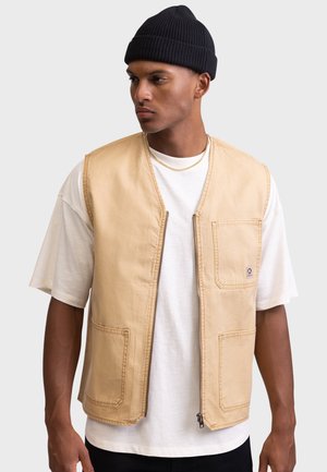 Gilet en coton beige avec deux poches avant et une fermeture zippée. Porté par-dessus un t-shirt blanc, avec un design minimaliste et une coupe décontractée.