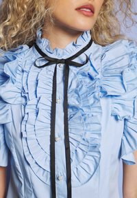 Kergelt sinisest volangiga pluusist, millel on pärlilukud ja kaelas must riba, tukkidega blond naine.