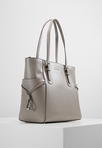 Sac fourre-tout gris en matériau texturé, avec deux poignées, fermeture zippée et poches latérales. Design minimaliste avec des détails matériels subtils.