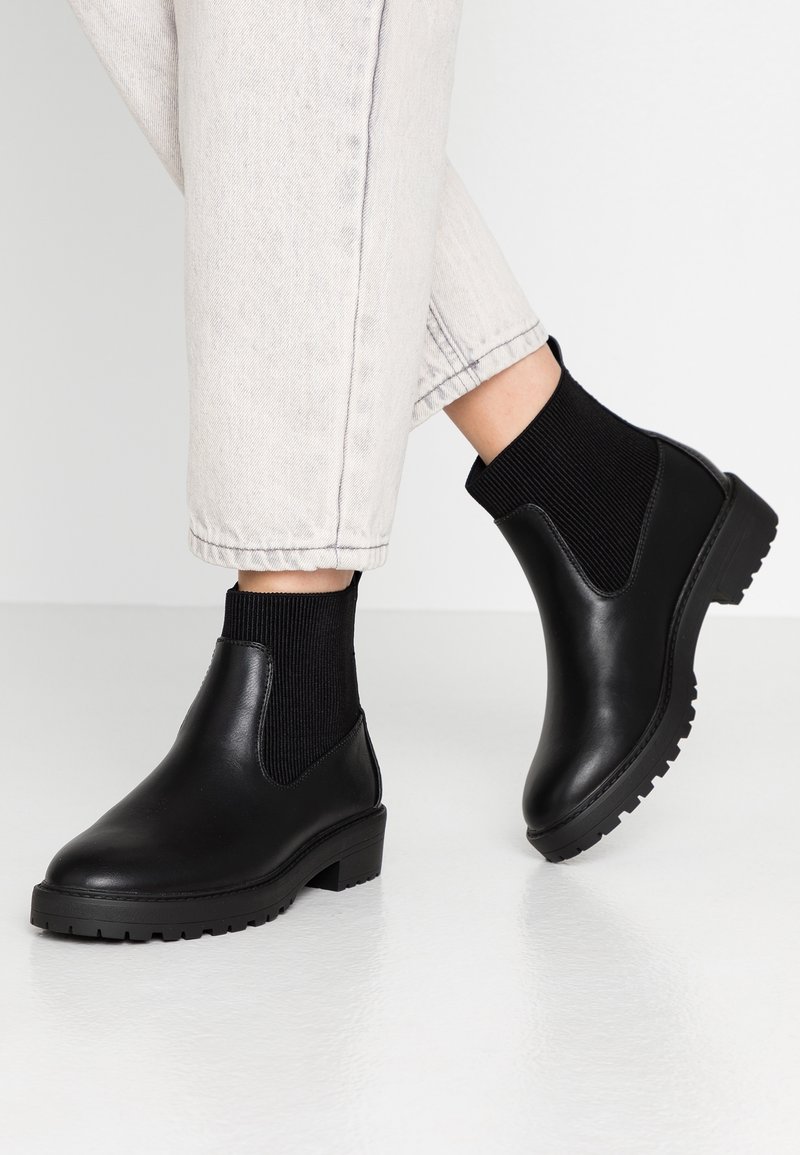 Personne portant un pantalon de couleur claire et des bottines Chelsea en cuir noir à semelles épaisses sur un sol blanc lisse.