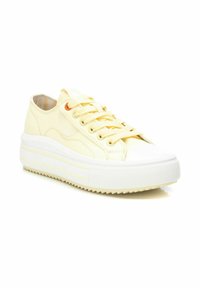 Zapatilla de lona amarilla con punta de goma blanca y suela, que presenta cordones planos, suela texturizada y detalle de costura lateral.