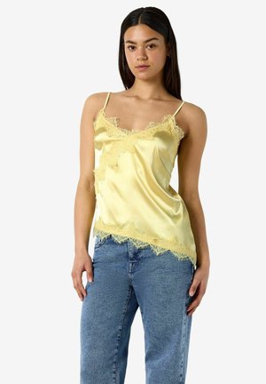 Noor naine, kes kannab kollast satiinsest kampsunit pitsilise äärisega ja siniseid teksapükse, seisab ühevärvilise valge tausta ees.