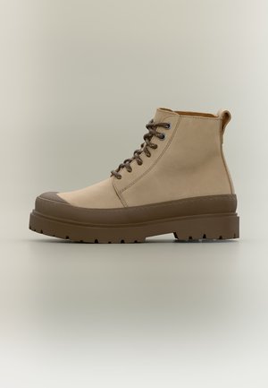 Bottine beige à la cheville avec semelle épaisse en caoutchouc marron, embout et lacets, avec une languette à l'arrière, présentée sur un fond uni.