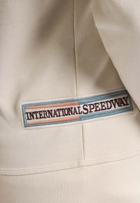 Tessuto color crema con una finitura testurizzata che presenta una toppa rettangolare. La toppa mostra la scritta "INTERNATIONALSPEEDWAY" in marrone, blu e pesca.
