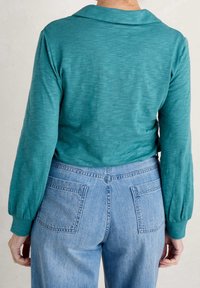 Maglietta lunghe maniche color teal con colletto e maniche leggermente a sbuffo, abbinata a jeans in denim a vita alta di colore blu chiaro con due tasche posteriori.