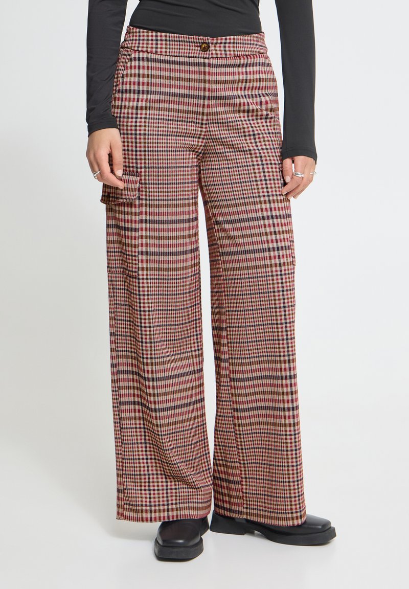 Personne portant un pantalon à carreaux à jambes larges avec une poche latérale, des chaussures noires et un haut noir ajusté à manches longues sur un fond uni.