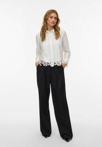 Femme portant un chemisier blanc à manches longues avec bordure en dentelle et un pantalon large noir, debout les mains dans les poches devant un fond blanc.