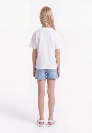 Niño con cabello largo y rubio que lleva una camiseta blanca, pantalones cortos de mezclilla azul claro y sandalias blancas, de pie de espaldas sobre un fondo blanco.