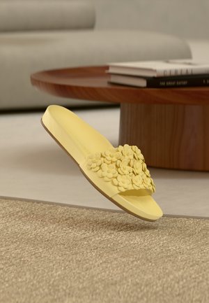 Yellow slide sandal made of smooth material, featuring a floral accent on the strap and a lightly textured sole.