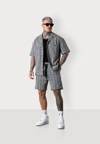 Kurzärmliges graues karierter Hemd und passende Shorts. Das Outfit bietet eine entspannte Passform, gewebte Textur und Seitentaschen. Tragen mit weißen Turnschuhen.