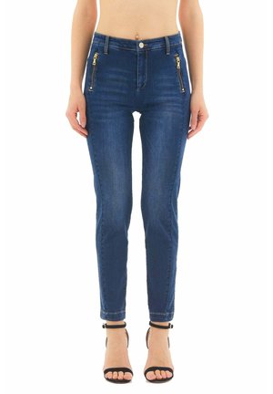 Woman wearing high-waist dark blue skinny jeans with front zipper pockets and black ankle-strap open-toe heels, standing against white background.