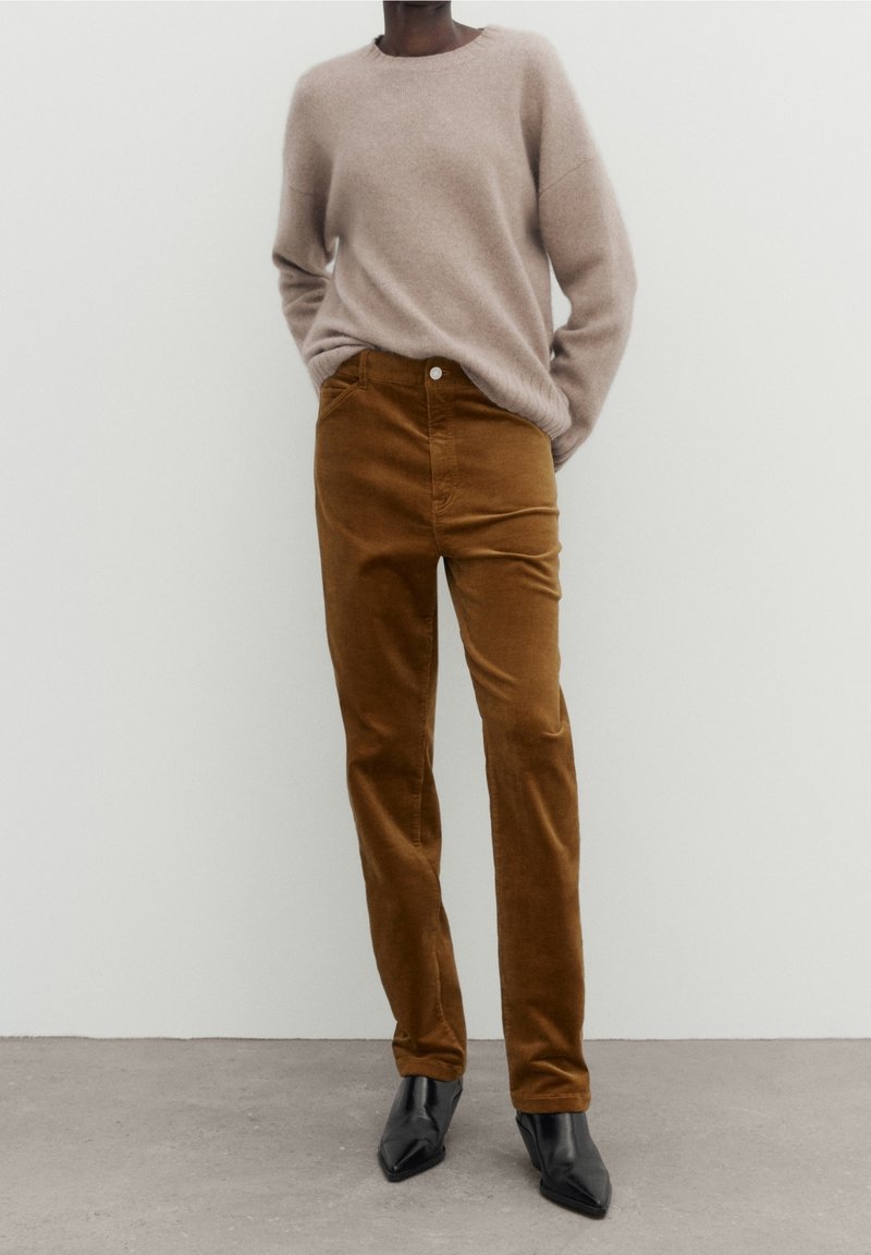 Pantalon en velours côtelé marron à la coupe ajustée, associé à un pull en tricot beige clair. Des talons noirs à bout pointu complètent la tenue.
