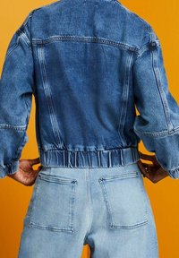 Blå denimjacka med en kortad design och elastisk midja, med långa ärmar och en blekt tvätt, ihop med ljusblå jeans.