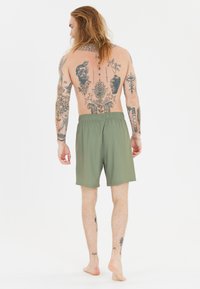 Gröna shorts med elastiskt midjeband, gjorda av lätt material. Figuren har en tatuerad rygg och är barfota.