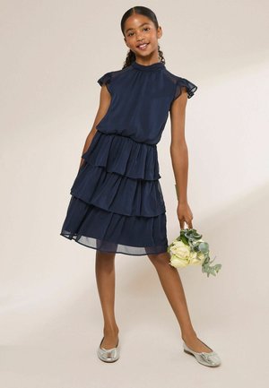 Mädchen trägt ein ärmelloses, marineblaues Stufenkleid und silberne Ballerinas, hält einen kleinen Strauß aus weißen Rosen und Grün.