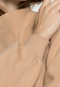 Světle béžový svetr s ribovanou texturou, nadýchanými rukávy a přiléhavými manžetami. Tkanina vypadá jemně a lehce s decentním leskem.