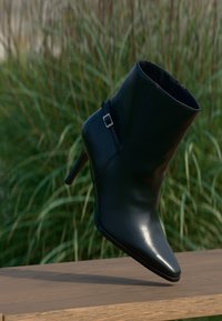 Black leather ankle boot with a pointed toe, high stiletto heel, and a silver buckle accent on the side, set against greenery.