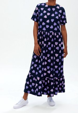 Osoba w granatowej sukni maxi z fioletowymi wzorami w serca i białych trampkach stoi na jednolitym tle.