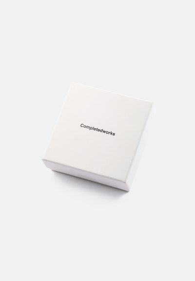 Λευκό τετράγωνο κουτί με λείο φινίρισμα. Στην κορυφή φέρει το κείμενο «Completedworks» σε απλή μαύρη γραμματοσειρά. Κλειστό και διακριτικό σχέδιο.