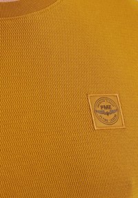 Gelbes texturiertes Strickshirt mit einem quadratischen Logo-Patch, auf dem "PME Flying Legend" und ein Emblem stehen. Rundhalsausschnitt; kurzes Mieder; lässiges Design.