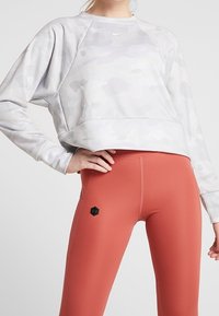 Felpa grigia chiara corta con un motivo camouflage e un piccolo logo, abbinata a leggings aderenti color ruggine con un accento esagonale.