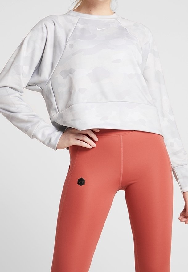 Felpa grigia chiara corta con un motivo camouflage e un piccolo logo, abbinata a leggings aderenti color ruggine con un accento esagonale.
