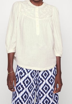 Lysebeige, tekstureret bluse med lange ærmer, knaplukning og blomsterbroderi på bærestykket, kombineret med mønstrede blå og hvide bukser.