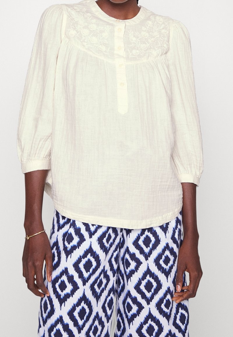 Blouse légère beige, texturée, manches longues, avec fermeture à boutons et broderie florale sur le yoke, associée à un pantalon bleu et blanc à motifs.