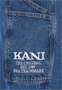 Tejido de mezclilla en azul oscuro con costuras naranjas; presenta un logotipo bordado en blanco "KANI" junto con el texto "THE ORIGINAL EST. 1989 REG. TRADEMARK."